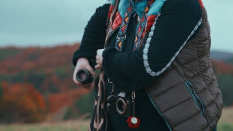 Close-up-shot-of-a-woman-changing-lenses-on-her-camera-with-beautiful-colourful-strap-with-orange-coloured-forest-in-the-background-during-a-windy-autumn-day-in-slow-motion