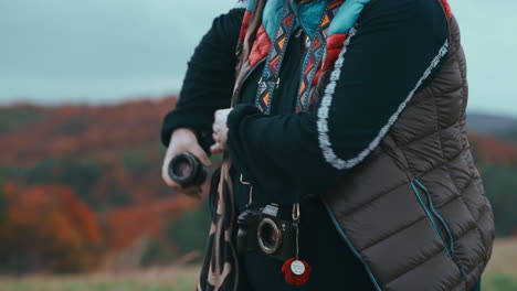 fotografía de cerca de una mujer cambiando las lentes de su cámara con una hermosa correa de colores con un bosque de color naranja en el fondo durante un ventoso día de otoño en cámara lenta