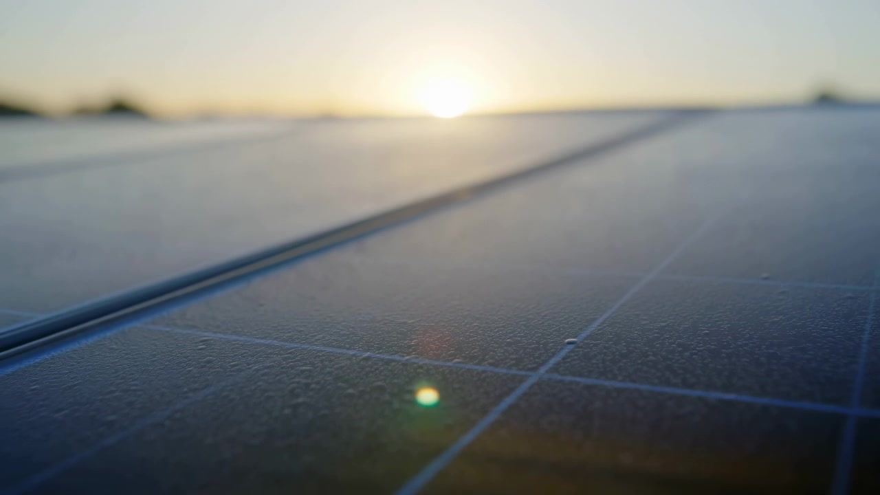 Primo piano tattile di una superficie fotovoltaica che mostra goccioline d'acqua, finitura scura e texturizzata, sottili linee a griglia blu e un morbido riflesso dell'alba su un dettaglio pulito e moderno di energia rinnovabile da molto in basso.