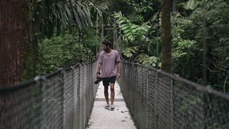 man walks across hanging bridge in rain forest with camera in hand, slow motion