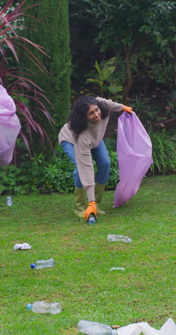 Vertical video: Woman seeing litter in yard, picking bottles and filling purple bag to clean yard