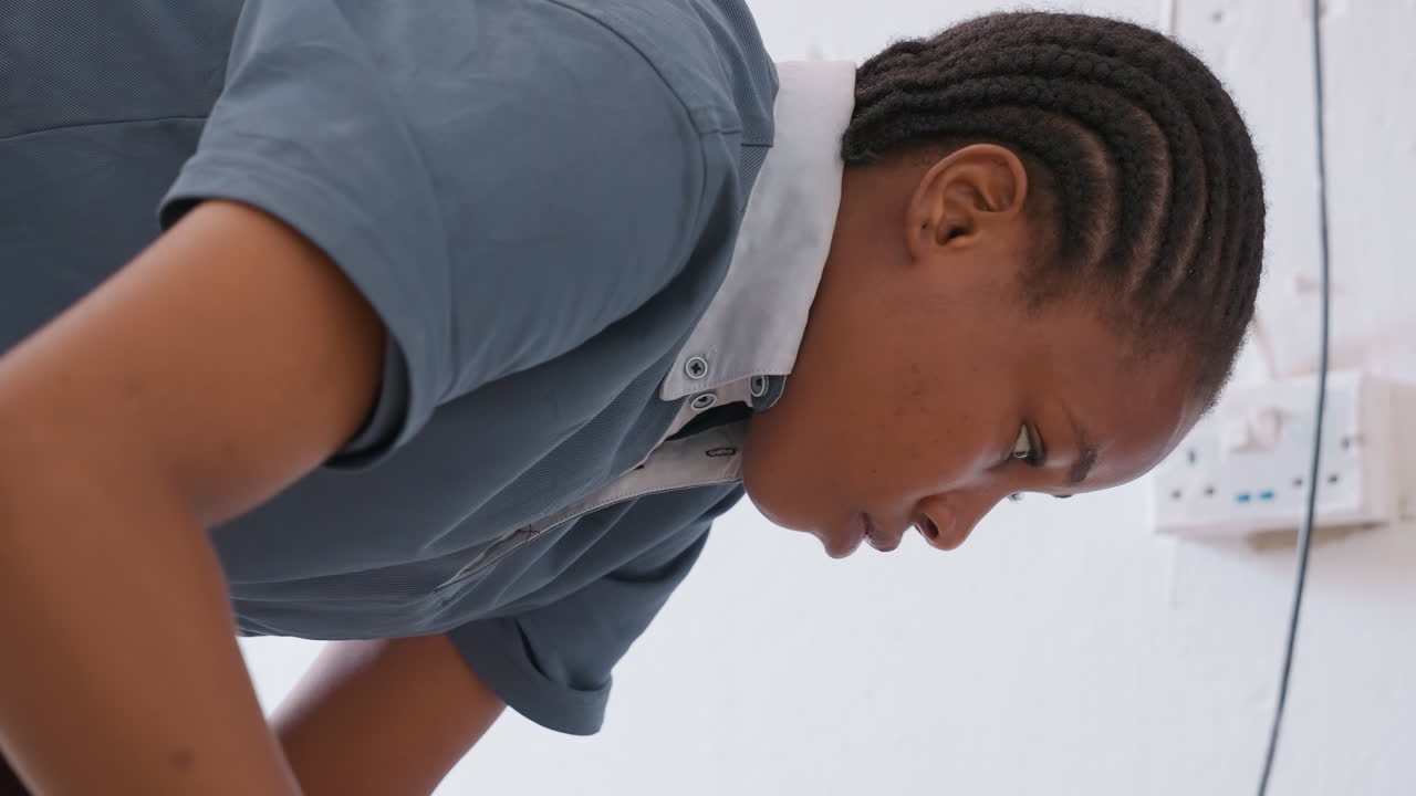 Black teenager sweeping house floor, focused. Braided hair in cornrows, wearing uniform shirt, leaning forward while sweeping with broom, careful posture and concentrated expression. Indoor white wall