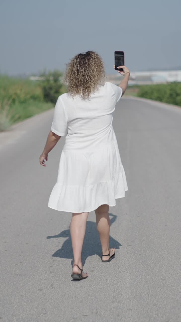 Donna con vestito bianco che cammina sulla strada e scatta una foto con il cellulare