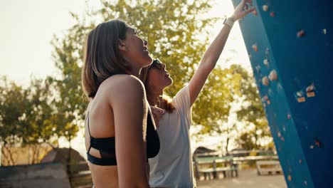 A-blonde-girl-in-black-glasses-and-a-white-T-shirt-tells-her-friend-Rock-girl-with-a-bob-hairstyle-in-a-black-top-how-they-will-climb-a-route-at-a-climbing-wall-on-a-sunny-day-in-the-summer