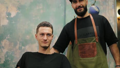 Der-Junge-Bärtige-Friseur-Lächelt-Und-Steht-Neben-Seinem-Zufriedenen-Kunden-Mit-Piercing-Im-Ohr-Und-Tätowierung,-Der-Auf-Dem-Stuhl-Sitzt