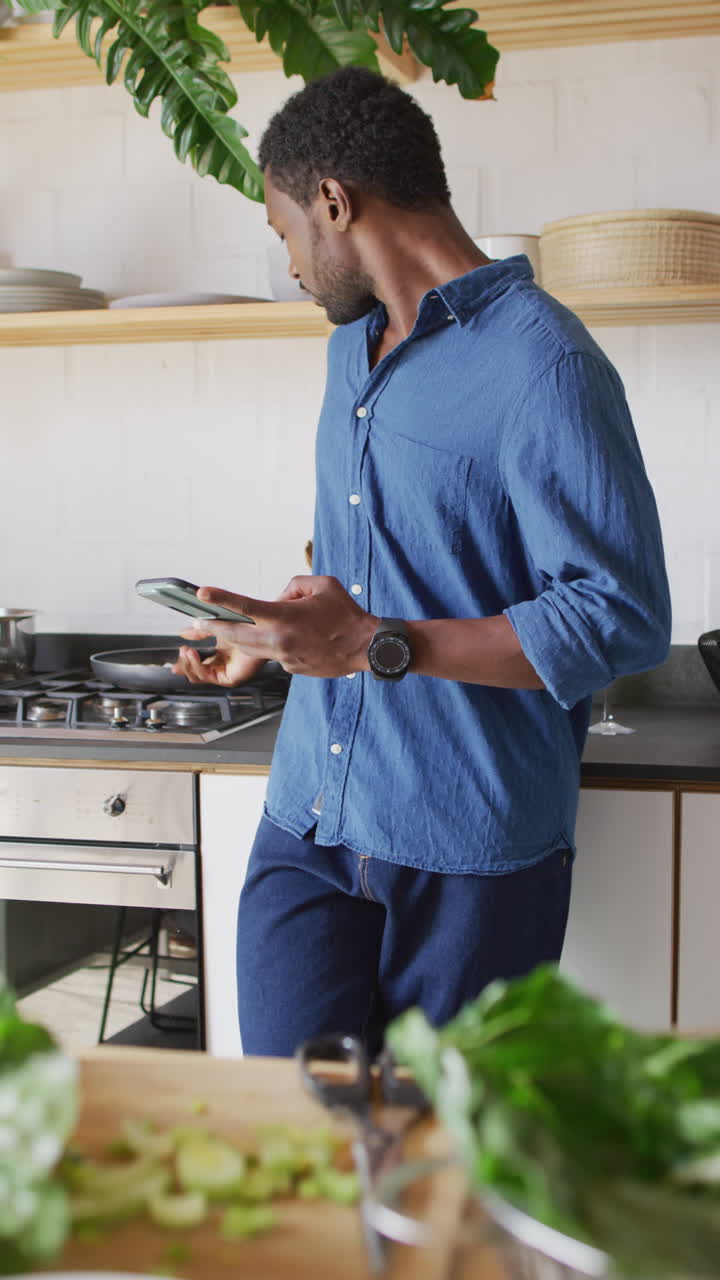 video vertical de un hombre afroamericano cocinando la cena en la cocina, usando un teléfono inteligente