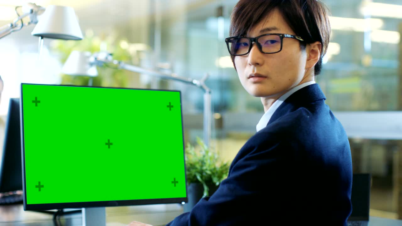 사무실에서 동아시아 사업가는 모의 녹색 화면을 가진 데스크 개인 컴퓨터에서 일합니다. 그는 극적으로 머리를 돌리고, 어깨를 통해 카메라로 바라보고, 동료가 사무실에 들어와 그녀의 책상에서 일어납니다.