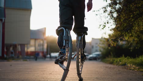 low back view of person walking on spring stilts during sunset wearing gray pants. warm evening sunlight creates long shadows, blurred trees and street in background emphasize motion, balance