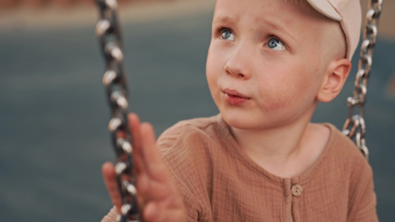 Young boy enjoys swinging at playground held gently by adult hand. Boy wears light colored cap and brown textured shirt as boy grasps chain swing. background is softly blurred emphasizing
