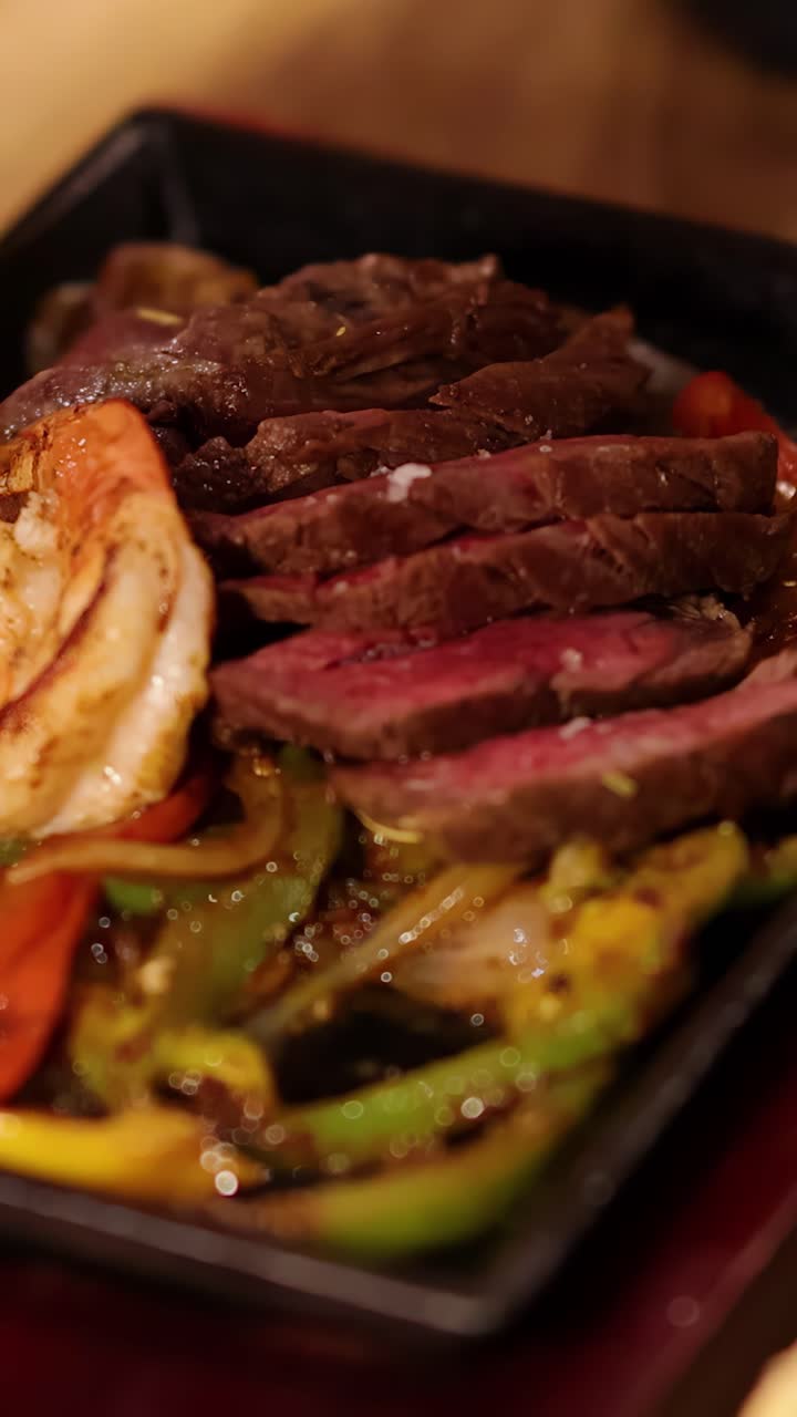 Primo piano di una forchetta che solleva gamberi e verdure da una padella calda con bistecca a fette, peperoni e cipolle, sotto una calda illuminazione da ristorante