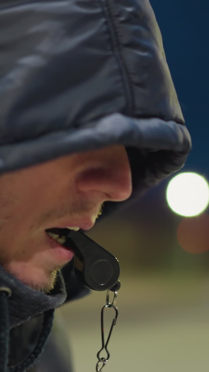 vista lateral de un hombre sentado con un silbato en la boca, con una chaqueta con capucha, el fondo está borroso y suavemente iluminado