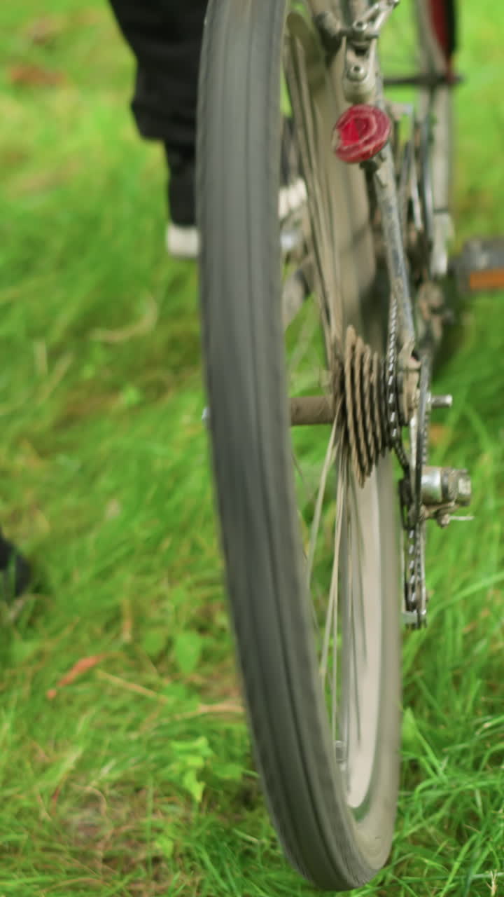 vista trasera de una bicicleta estacionada en un campo de hierba, alguien con pantalones negros se acerca la persona levanta la parte trasera de la bicicleta y los pedales, haciendo que los neumáticos giren, con un enfoque en los neumáticos