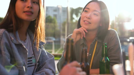 group of three japanese friends talking and holding beers while sitting at table outdoors in a sunny day