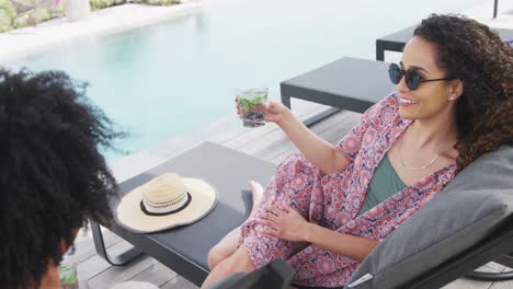 video of happy diverse couple relaxing with drinks and talking by swimming pool at holiday home