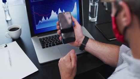 caucasian man wearing face mask holding smartphone while sitting on his desk at modern office