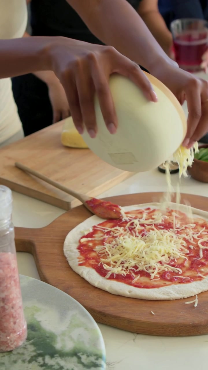 Video verticale: Una cuoca in vestito color crema spalma la salsa e aggiunge formaggio alla pizza al bancone con gli amici.