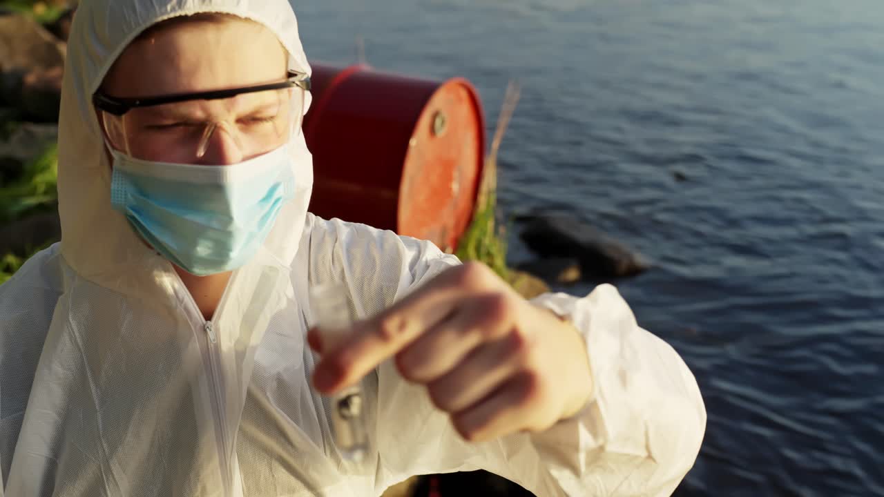 Giovane ispettore ambientale o scienziato caucasico in tuta protettiva e mascherina che tiene in mano un campione d'acqua in una provetta mentre analizza l'inquinamento vicino a un fiume con barili arrugginiti, carrellata