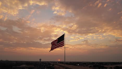 Large-American-flag-waves-patriotically-in-the-wind-above-rising-sun-and-empty-serene-cloudy-sky