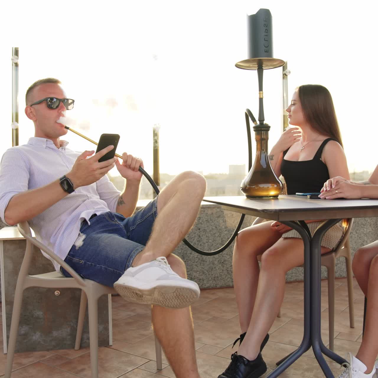 Company of young people having good time in the café outdoors. Man smoking hookah looking at phone and chatting with girls