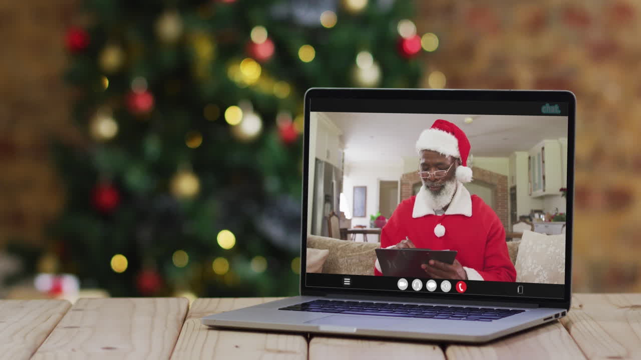 hombre afroamericano senior en disfraz de santa en videollamada en computadora portátil, con árbol de navidad