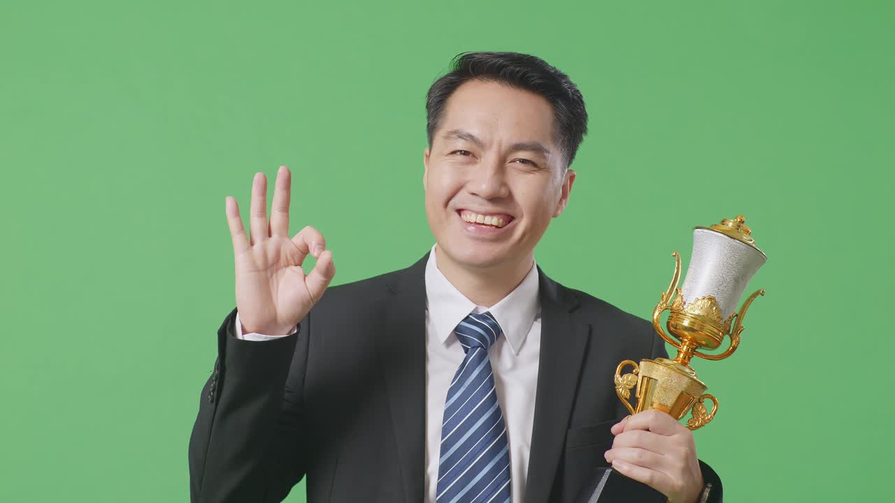 슈트와 타이를 입은 아시아 사업가, 금메달과 트로피를 들고 오케이 제스처를 보여주며 카메라에 미소 짓고, 스튜디오의 녹색 스크린 배경에서 첫 번째 우승자로