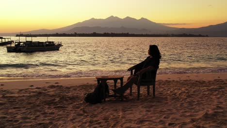 Chica-Sentada-Sola-En-Una-Tranquila-Playa-Exótica-Sin-Gente,-Viendo-Una-Hermosa-Laguna-Con-Barcos-De-Pesca-Al-Atardecer,-Indonesia