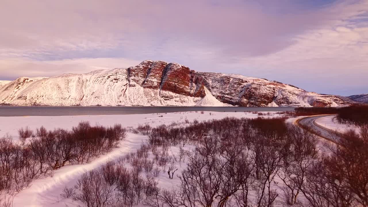 Aerial drone footage slowly flying over gorgeous leafless nature landscape during sunset or sunrise in Norway with Giemaš or Giemas mountain is in the background. A lonely road is on the right also.