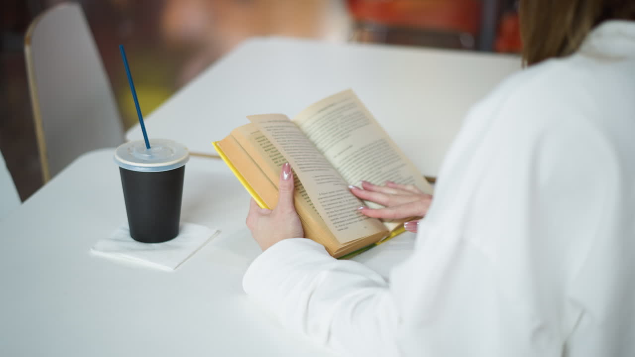vista trasera de una mujer sentada en una mesa hojeando un libro con una taza de café a su lado en la mesa, tranquila y enfocada en un entorno comercial, con fondo suave y colores vibrantes