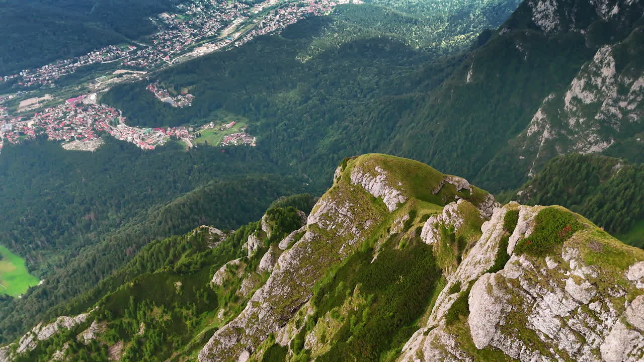 Flight over the mossy rocks of the huge mountain. View on the residential area and green woods at the mountain foot. The Bucegi Mountains of the Southern Carpathians, Romania