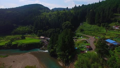 Die-Luftaufnahme-Bewegt-Sich-über-Einen-Ruhigen-Fluss-Und-Zeigt-Ein-Malerisches-Japanisches-Dorf-In-Einem-Grünen-Tal