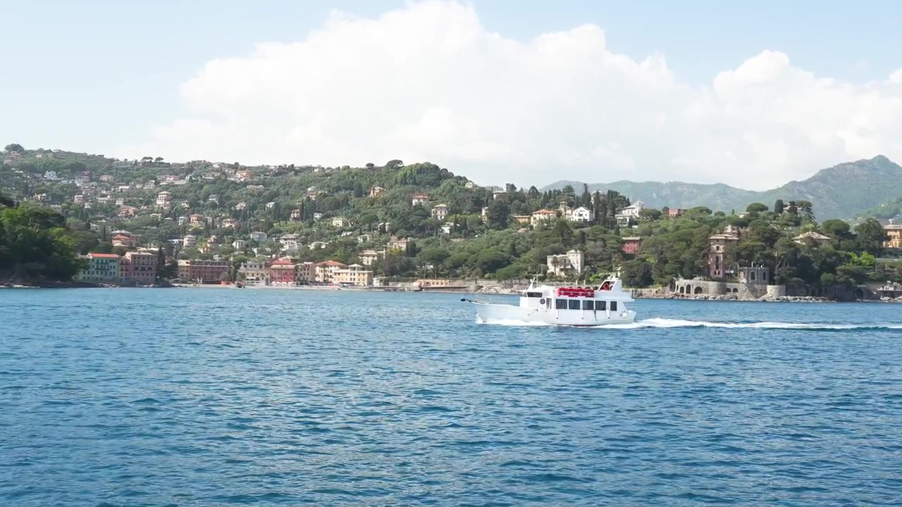Un'ampia inquadratura serena mostra una pittoresca città costiera con ville collinari, acqua cristallina e una barca passeggeri bianca che scivola sull'acqua in pieno giorno, con nuvole leggere in cielo.
