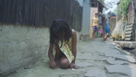 young indian poor orphan girl, sitting on ground playing with small rock on ground and soil, rural area