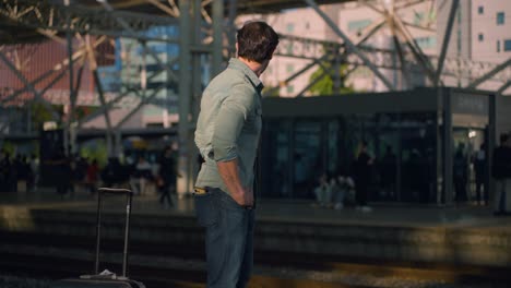 stylish traveler checking for train and turning around at seoul station, korea, solo male in urban transport hub with crowds behind on other side of tracks