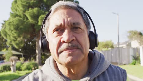 Video-close-up-of-senior-biracial-man-in-sports-clothes-and-headphones-running-on-sunny-street
