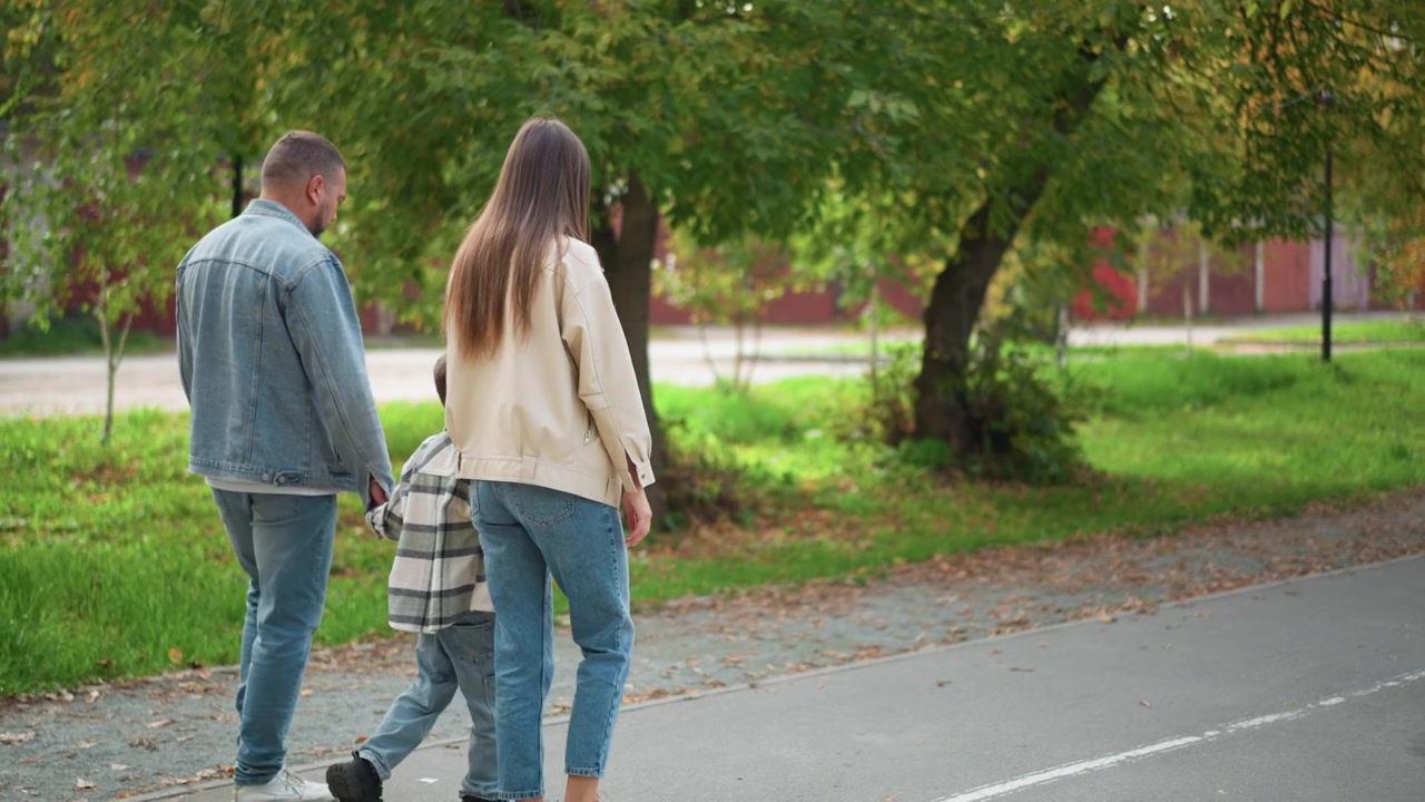 Familia de tres caminando juntos por un sendero pavimentado a través de un parque verde con árboles y césped. Hombre con chaqueta de mezclilla, mujer con top beige, y un niño pequeño tomados de la mano entre ellos, todos vestidos de forma casual.