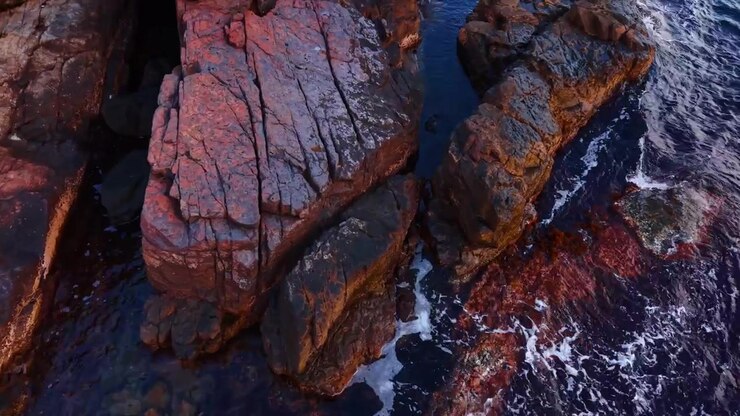 Luce calda su formazioni rocciose costiere bagnate con mare scuro. Vista aerea ravvicinata di massi stratificati marroni e rossi tra acqua blu scuro con schiuma bianca delle onde.