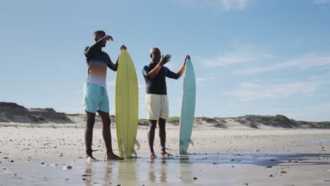 padre afroamericano y hijo adolescente de pie en una playa sosteniendo tablas de surf y hablando