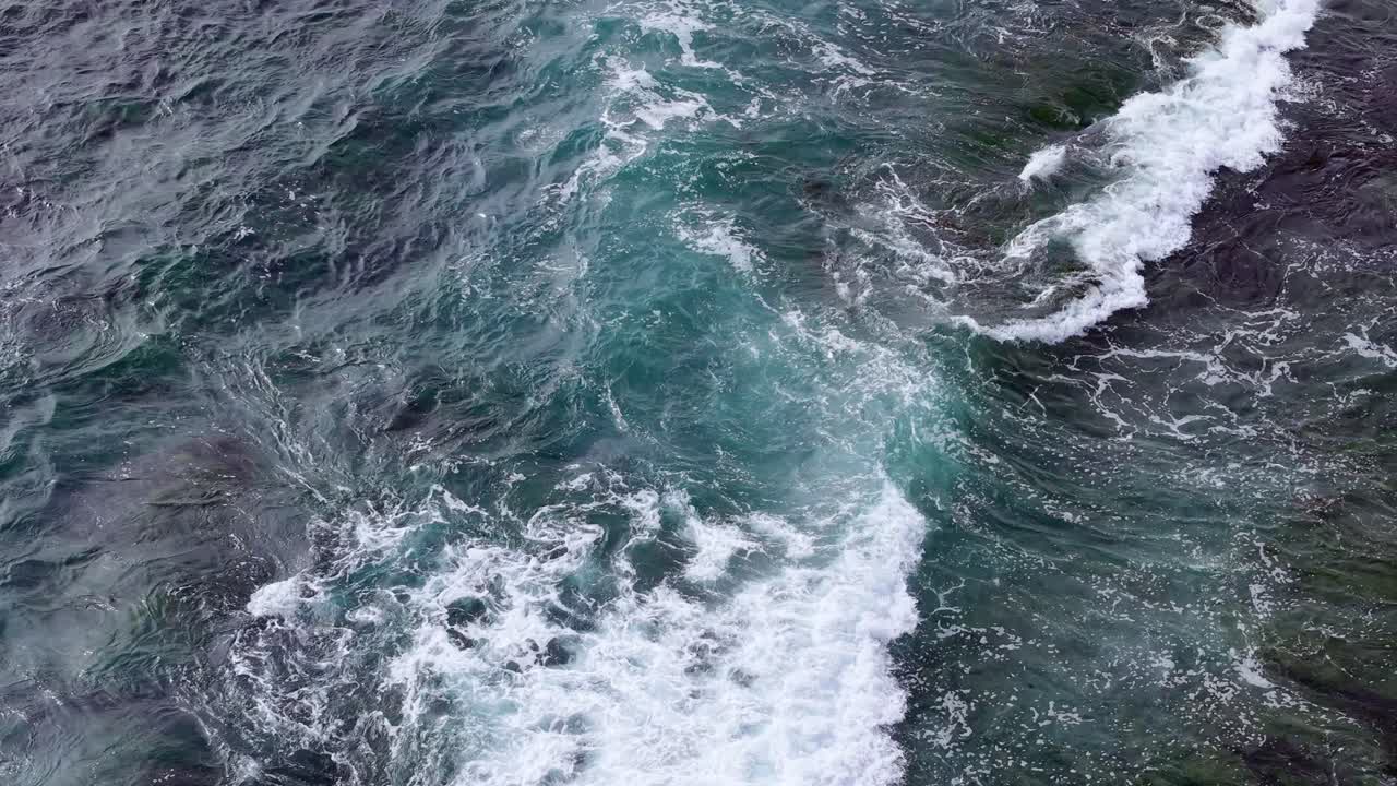 Вид сверху с дрона на волны, накатывающие на неглубокий скалистый риф в бирюзовых прибрежных водах. Чистый океан обнажает подводную текстуру, когда белая вода сталкивается с краем рифа