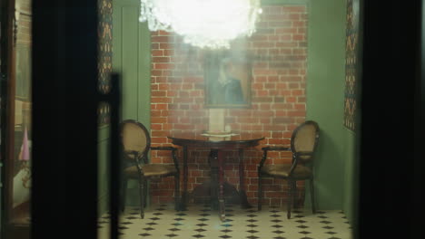 view of a hand closing a glass door to a cozy room with two chairs and a table. shot with a handheld camera, capturing the room's inviting ambiance with a chandelier, artwork, and vintage decor