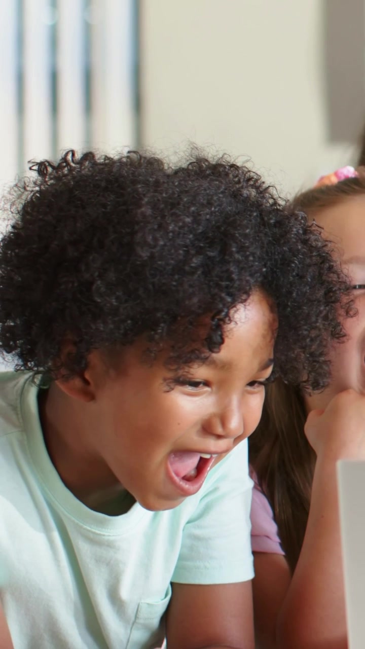 Vertical video: Leaning boy shifting into laughter watching laptop at home, mint tee, girl smiling