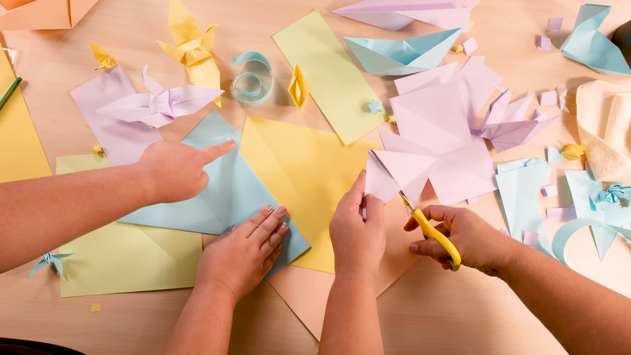 Two people fold and cut pastel paper for origami, overhead view, warm indoor lighting