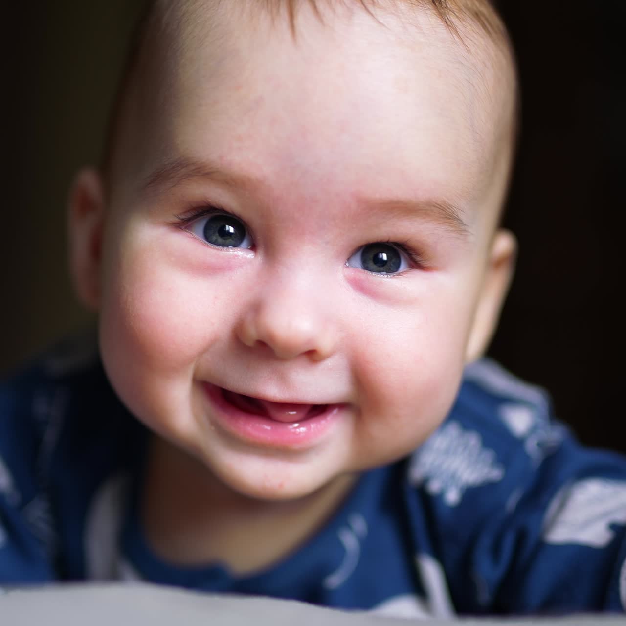 Beautiful grey-eyed child in blue clothes smiles to the camera. Baby boy lies on bed and moves cheerfully with his feet. Close up