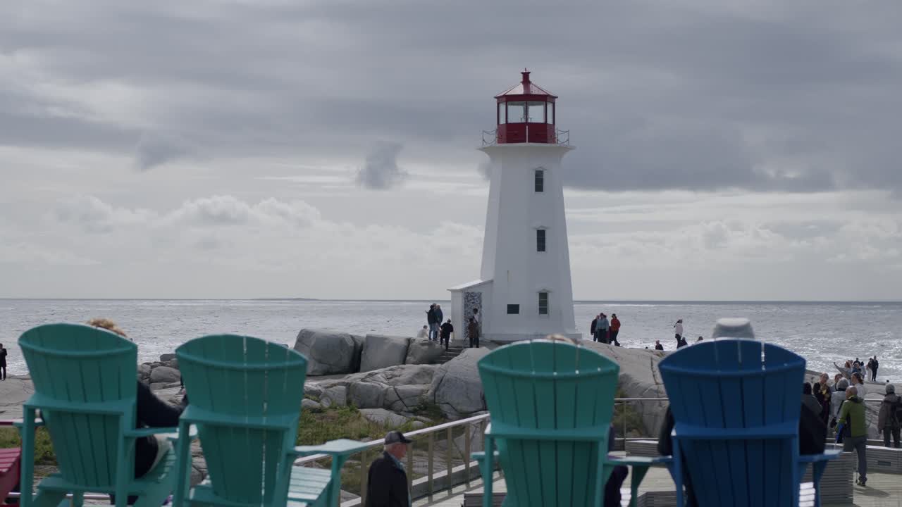 Tourists walking by Peggy's Cove lighthouse with Adirondack chairs in the foreground