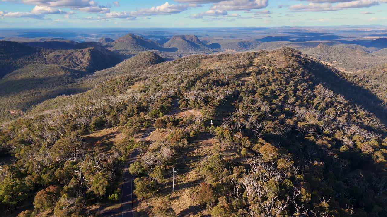 Drone camera glides above a rugged, forested mountain ridge in Warrumbungle National Park, New South Wales, under warm sunset lighting with expansive landscape views