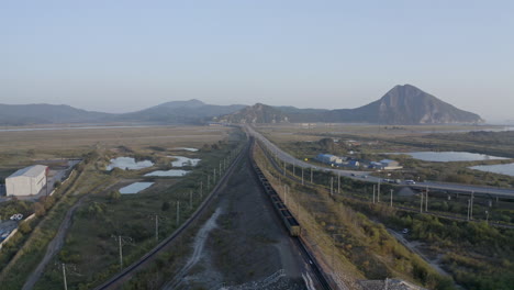 slow descent aerial shot of long full loaded coal cargo train carriage moving through the fields with mountains in the background on the sunset, russian federation