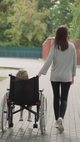 mother helps little daughter to move in wheelchair on path in city park. woman and girl with spinal cord injury enjoy walk on spring day backside view