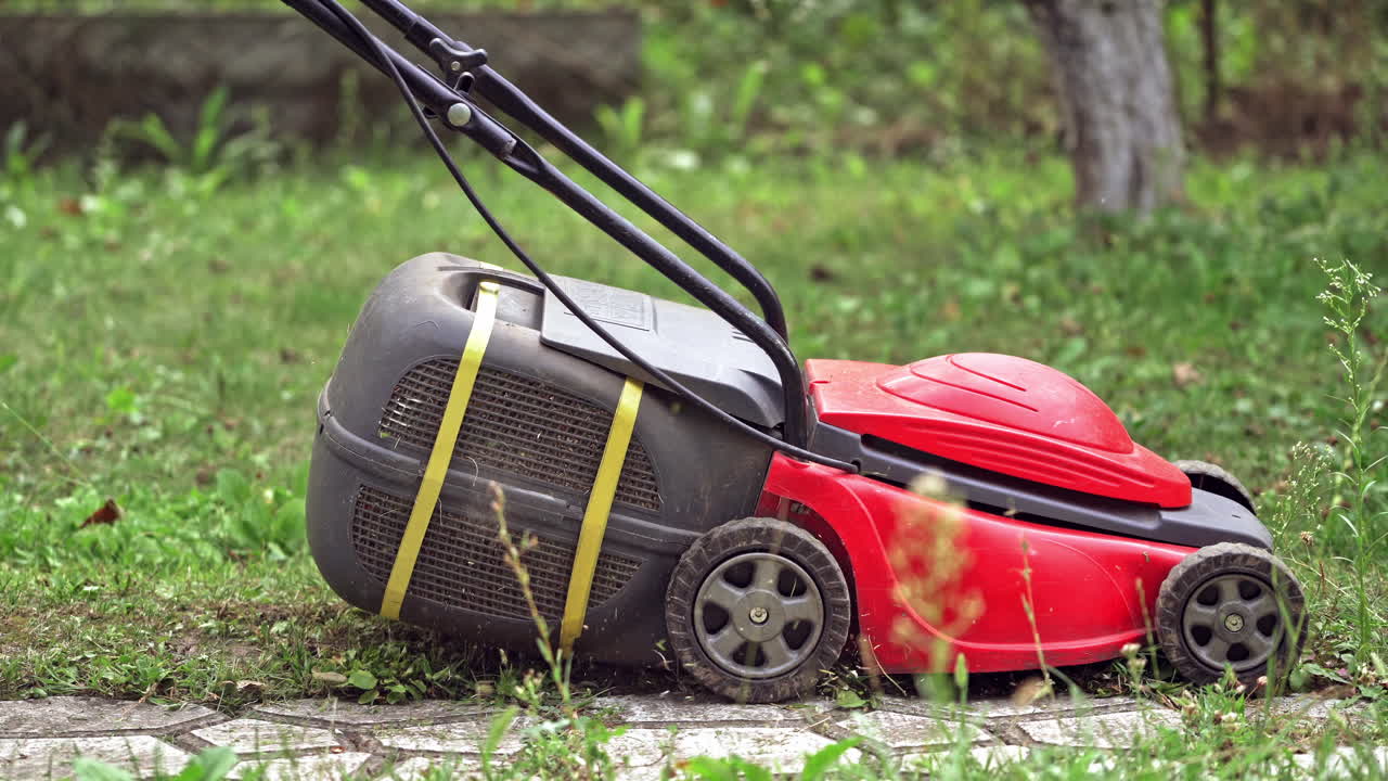 Modern technology of cutting grass in the garden. Red lawn mower cutting grass in the backyard. Corded electric lawn machine outdoors.