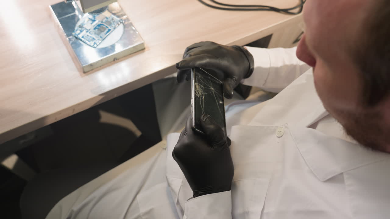 A downward view of a technician in a white lab coat and black gloves examining a cracked smartphone screen
