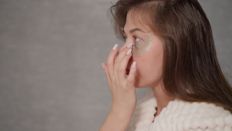 lady in plush robe applies moisturizing patch under eye in light place. pretty lady has beauty day enjoying cosmetics to moisturize skin in shower