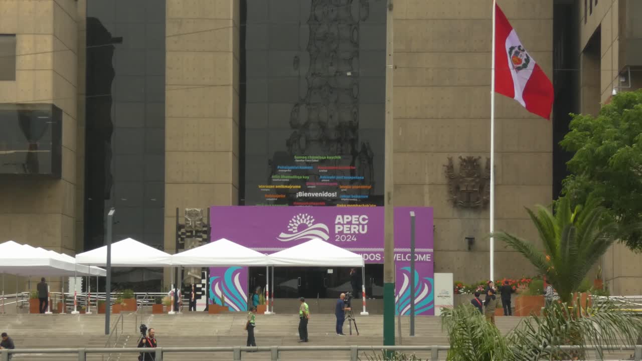 Entrance for the APEC 2024 summit in Lima, Peru. A few people walk around and a Peruvian flag blows in the wind on a tall flag post.